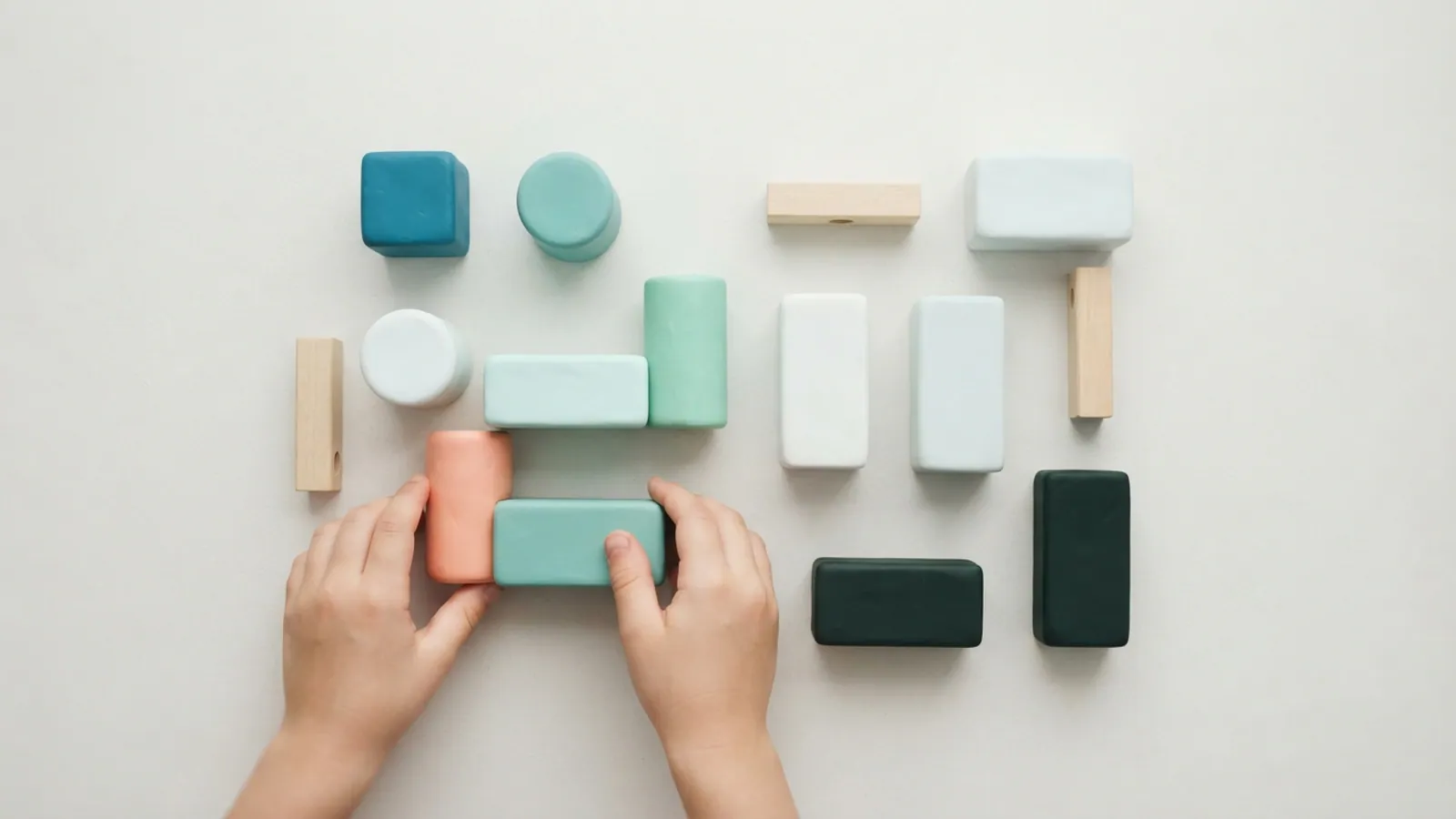 An overhead flat lay of plain building blocks and connectors on a light surface, with a child's hands arranging them.